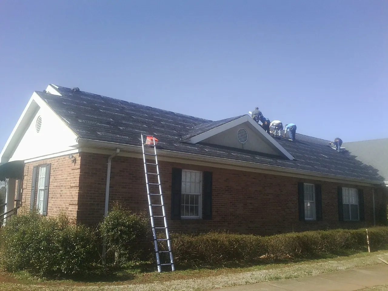 Crew tearing shingles off a brick ranch roof with a ladder set — Metal Roof Repair work by our crew in Rocky Ford
