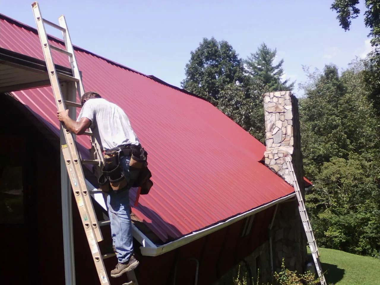 Roofer on a ladder beside a red metal roof and stone chimney — Soffit & Fascia Repair by Roofing Brothers in Rocky Ford